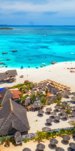 Aerial view of tropical sandy beach with palms and umbrellas at sunny day. Summer holiday. Landscape with palm trees, hotels, buildings, white sand, boats and yachts in azure sea. View from above